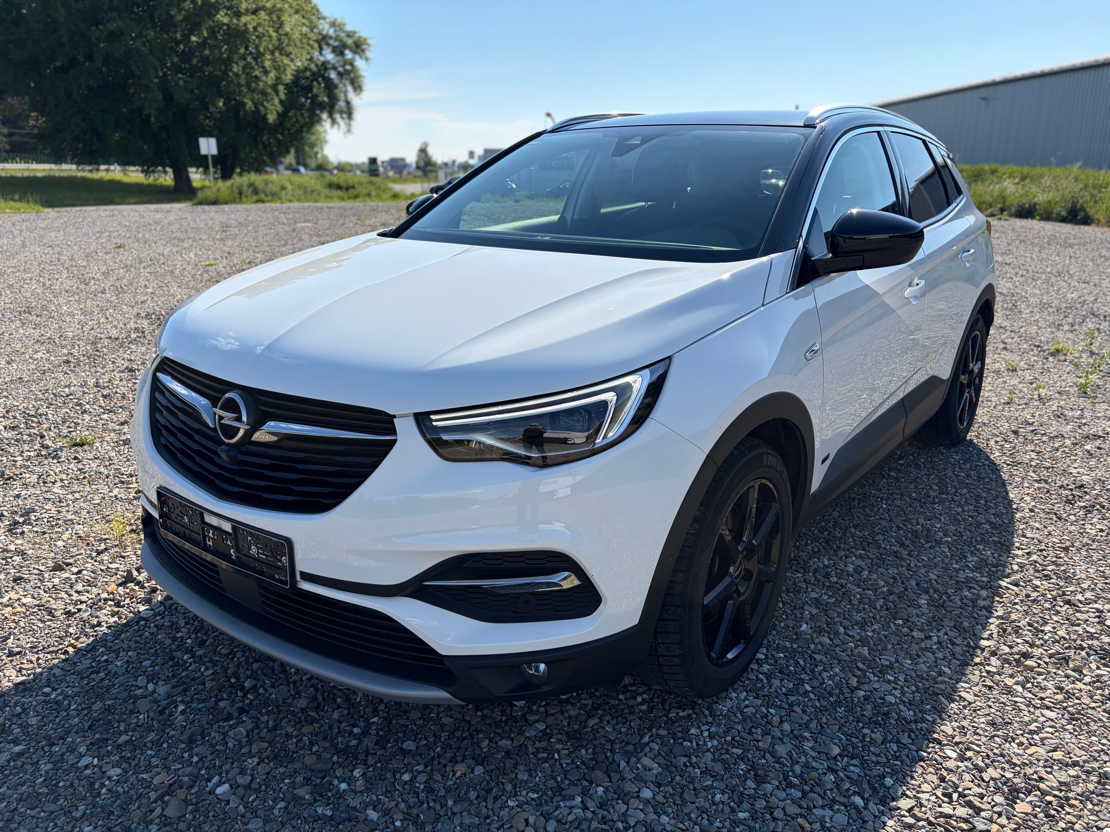 Ein weißer Opel Grandland SUV mit schwarzen Felgen steht auf einem sonnigen Schotterparkplatz.